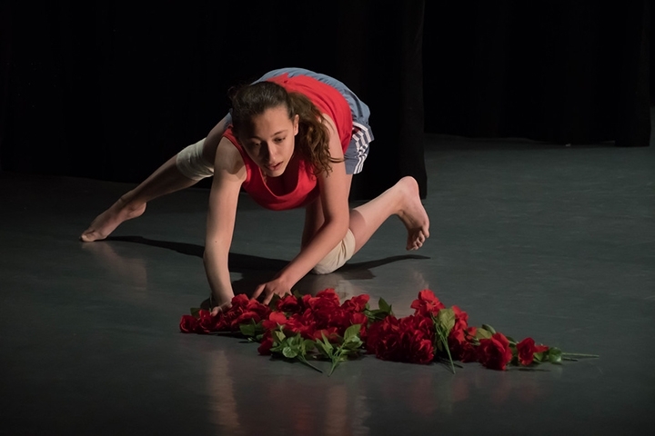 A single dancer in a crouch, reaches toward a pile of roses on the stage floor.