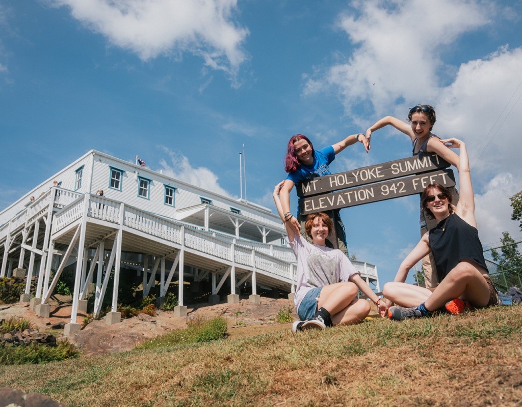 Mountain Day 2024 - Four students make a heart shape with their arms around the Mt. Holyoke state park sign at the top of the mountain.