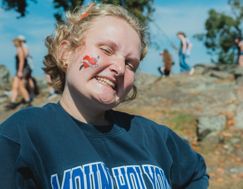 Mountain Day 2024 - Student wearing a Mount Holyoke sweatshirt smiles with a temporary tattoo on their cheek.