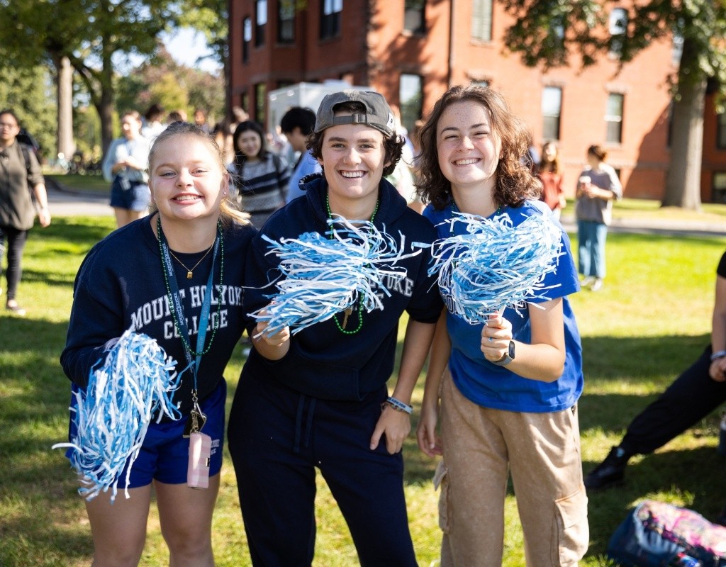 Three students together on Pageant Green holding blue and white pom poms