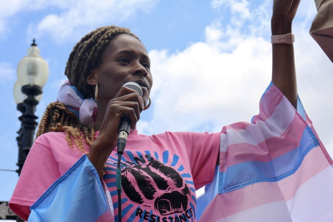 This is a photograph of Chastity Bowick at a demonstration; she is standing holding a microphone in one hand, the other arm raised. She wears a flag of pink, blue and white stripes, the colors of transgender pride. The blue sky backdrop matches the blue stripe in the flag.