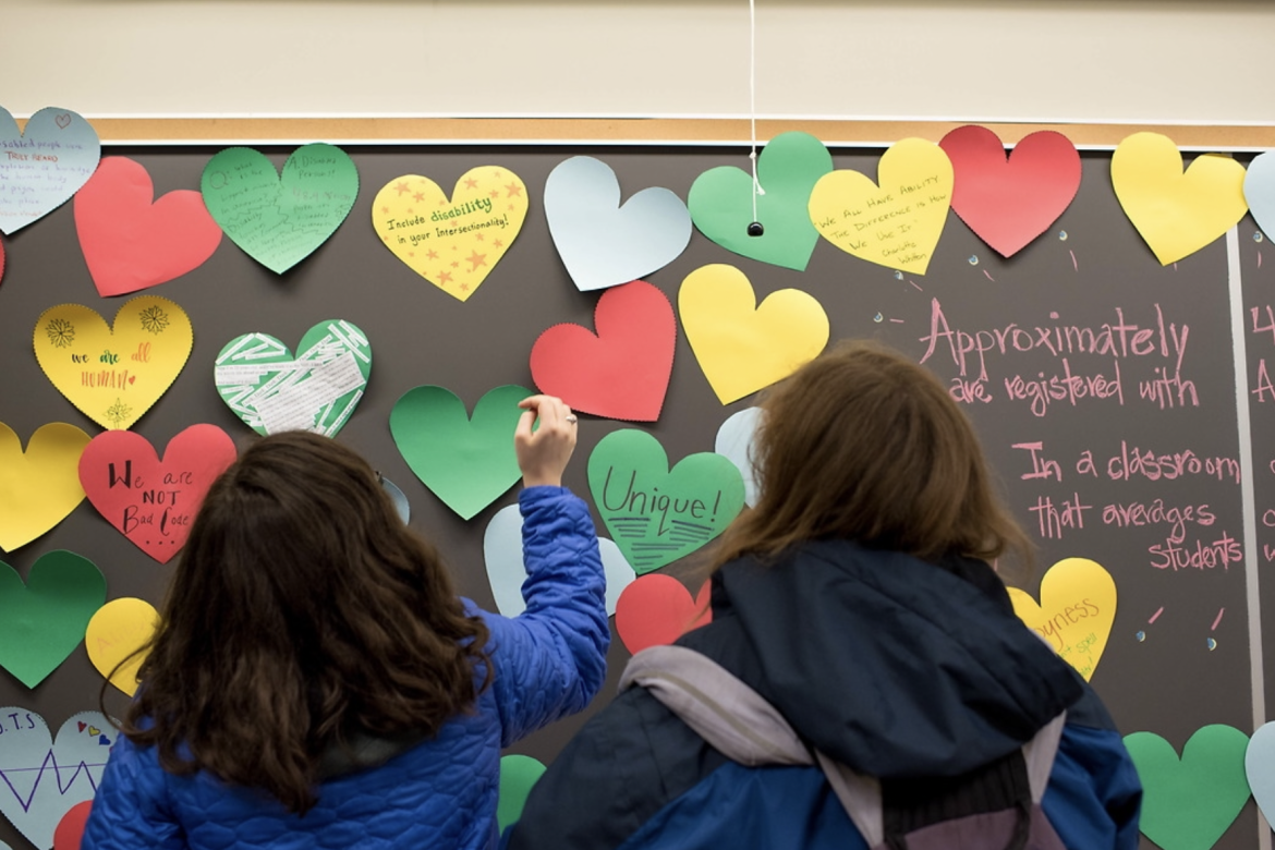 Students standing at a blackboard covered with stickynote hearts containing messages about disability