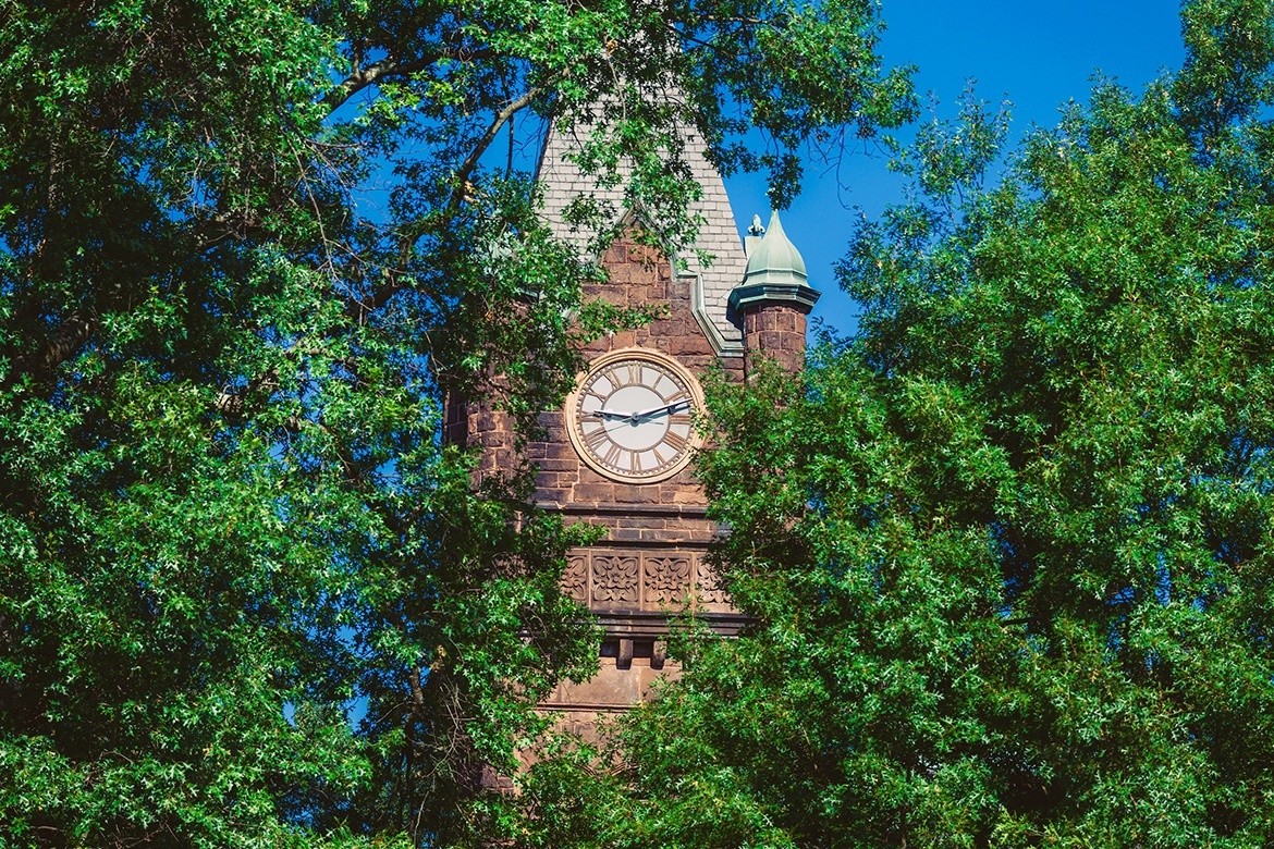 Mount Holyoke College's Clocktower slightly obscured by green leaves with a bright blue sky.