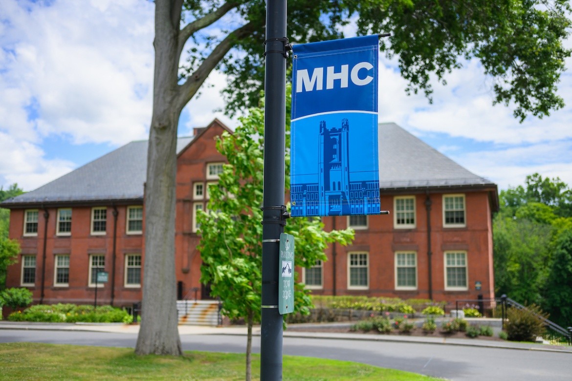Mount Holyoke campus - a banner on a sign with MHC and the clock tower. Photo by Max Wilhelm, 2024.