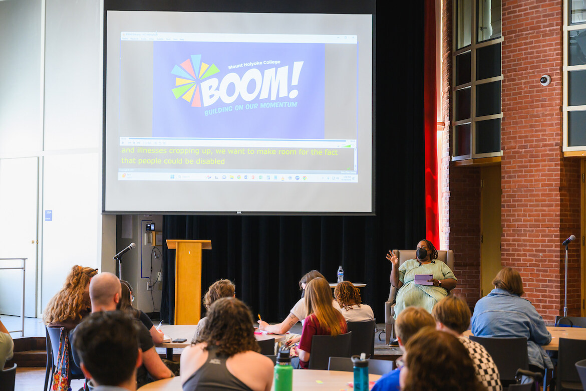 Audience sits at tables in the Great Room, BOOM presentation in the background.