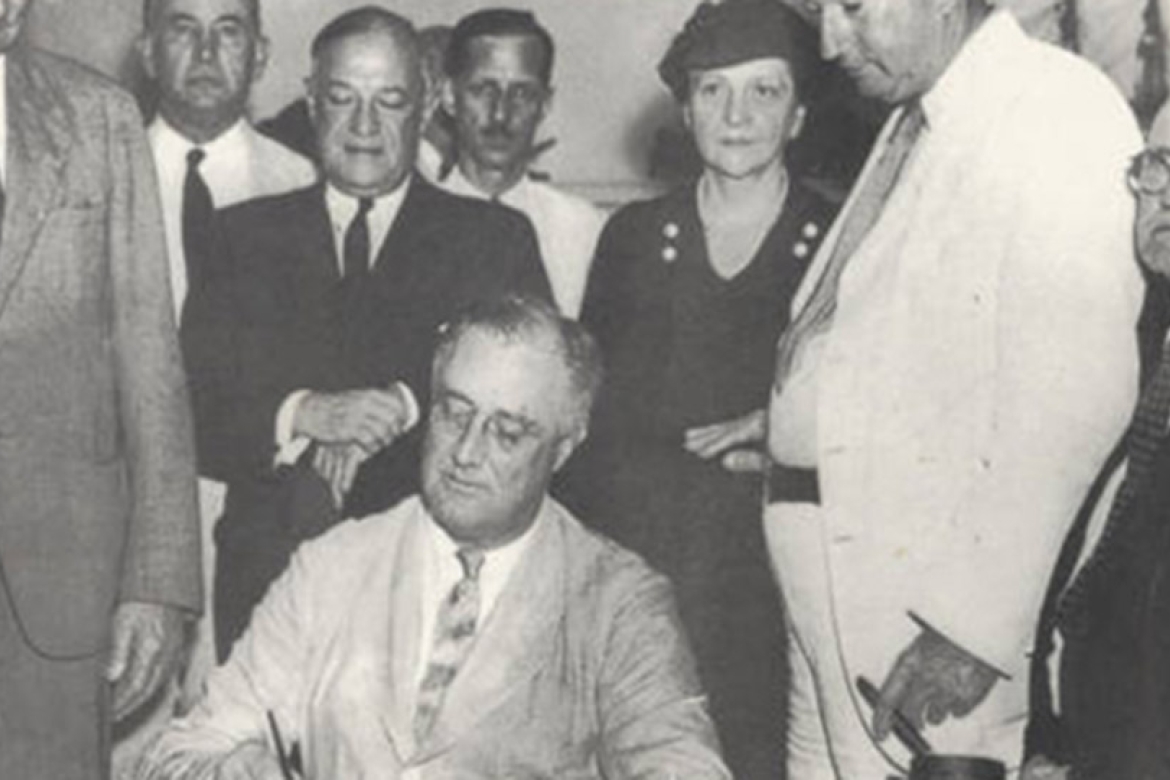 Frances Perkins looks on as President FDR signs the Social Security Act into law.