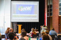 Audience sits at tables in the Great Room, BOOM presentation in the background.