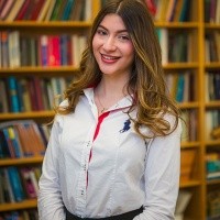 Alexandra Mihailopol ’26 in a room with bookshelves filled with books behind her. She has long brown hair, wearing a white shirt with a bit of red. She is smiling.