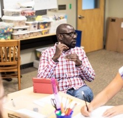Professional and Graduate student in a classroom, explaining something and gesturing while others take notes