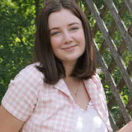 Brigid Mullen ’25, wearing a pink and white checked jacket over a floral top, stands in front of a wooden fence and greenery and smiles at the camera.