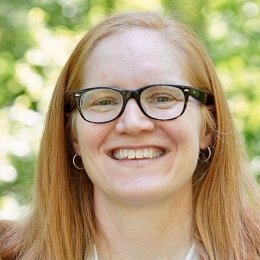 Andrea Ricketts-Preston, Athletic Director. A smiling woman with strawberry blonde hair, dark glasses, wearing a white shirt and a dark blue jacket in front of greenery.