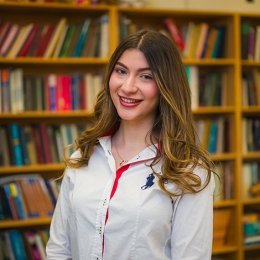 Alexandra Mihailopol ’26 in a room with bookshelves filled with books behind her. She has long brown hair, wearing a white shirt with a bit of red. She is smiling.