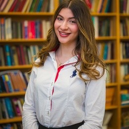 Alexandra Mihailopol ’26 in a room with bookshelves filled with books behind her. She has long brown hair, wearing a white shirt with a bit of red. She is smiling.