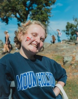 Mountain Day 2024 - Student wearing a Mount Holyoke sweatshirt smiles with a temporary tattoo on their cheek.