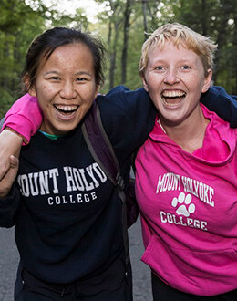 Two students wearing Mount Holyoke sweatshirts in a wooded area.