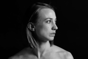 Ellie Goudie-Averill - a black and white photo of a woman with long hair looking to the right, a black background behind her, her face and shoulder lit by a light off screen.
