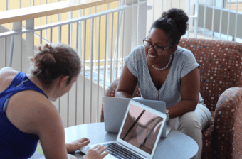 Professional and Graduate students sitting across from one another with laptops, one is smiling the other is engaging.