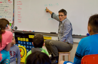 A teacher in front of a class, pointing at math on a white board.