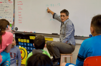 A teacher in front of a class, pointing at math on a white board.