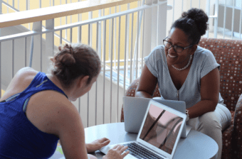 Professional and Graduate students sitting across from one another with laptops, one is smiling the other is engaging.