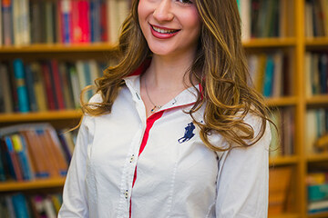 Alexandra Mihailopol ’26 in a room with bookshelves filled with books behind her. She has long brown hair, wearing a white shirt with a bit of red. She is smiling.