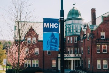 Banner on a light pole in front of Safford Hall with a newly blooming tree in the background. Mount Holyoke campus, April 2025. Photo by Max Wilhelm.