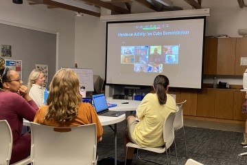 Several students listen to a lecture in a classroom. A teacher stands at a podium, and a screen shows faces of online students from across the state.