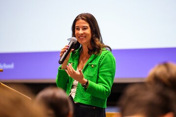 Caroline Nobo ’07, wearing a bright green jacket and holding a microphone, speaks in front of an audience.