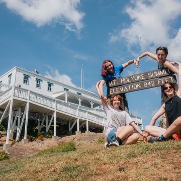 Mountain Day 2024 - Four students make a heart shape with their arms around the Mt. Holyoke state park sign at the top of the mountain.