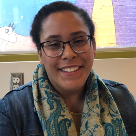 Stephani Lopez Rodriguez, MAT’19, smiling in front of a screen wearing a blue jacket and blue and cream scarf