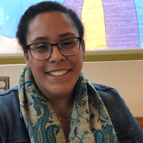 Stephani Lopez Rodriguez, MAT’19, smiling in front of a screen wearing a blue jacket and blue and cream scarf