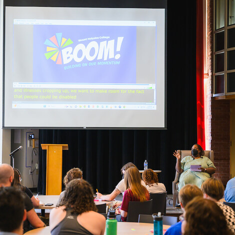 Audience sits at tables in the Great Room, BOOM presentation in the background.
