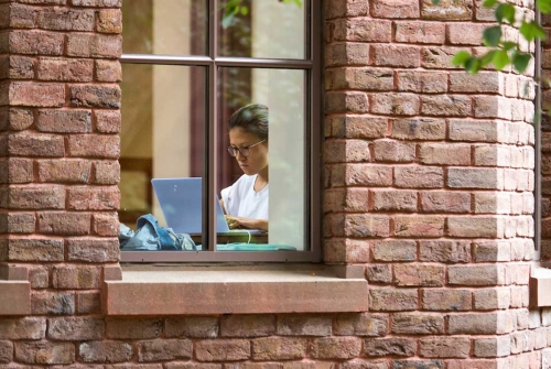 A student working on a laptop in the llibrary as seen through a window outside the building