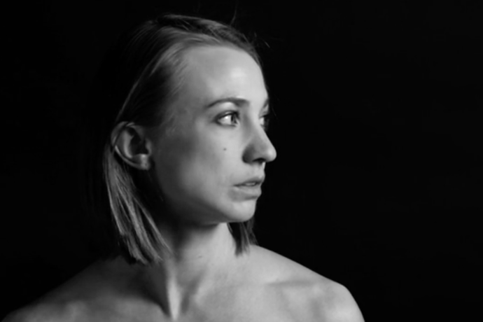 Ellie Goudie-Averill - a black and white photo of a woman with long hair looking to the right, a black background behind her, her face and shoulder lit by a light off screen.