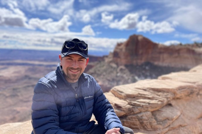 Ben Boatwright (image description: a while man wearing shades of blue with a mustache and small beard wearing a hat with a mountain and blue cloudy sky behind him)