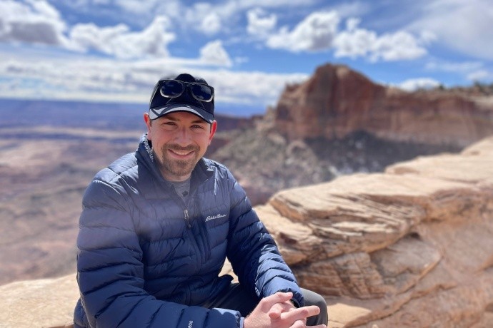 Ben Boatwright (image description: a while man wearing shades of blue with a mustache and small beard wearing a hat with a mountain and blue cloudy sky behind him)