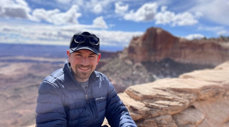 Ben Boatwright (image description: a while man wearing shades of blue with a mustache and small beard wearing a hat with a mountain and blue cloudy sky behind him)