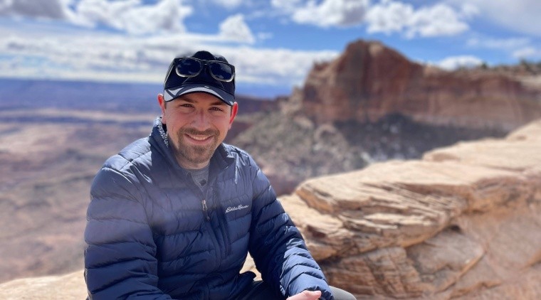 Ben Boatwright (image description: a while man wearing shades of blue with a mustache and small beard wearing a hat with a mountain and blue cloudy sky behind him)