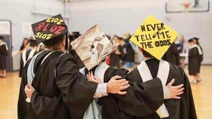 This is a picture of the back of three graduates wearing decorated mortarboards. The left one reads, "2019." The center one is decorated with a bee in pink, light blue and white. The one on the right reads "Never tell me the odds."