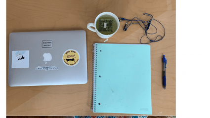 A closed laptop with stickers on the cover, a cup of tea, a notebook, pen and a headphones lying on a table