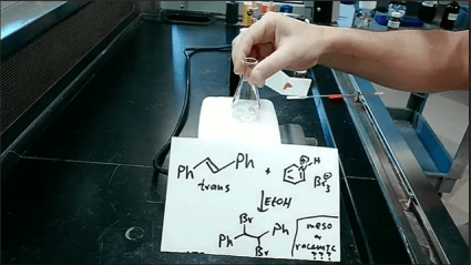 This is a still image from one of McMenimen's teaching videos, showing a hand grasping a small flask over a lab bench, with chemistry notations in the foreground.