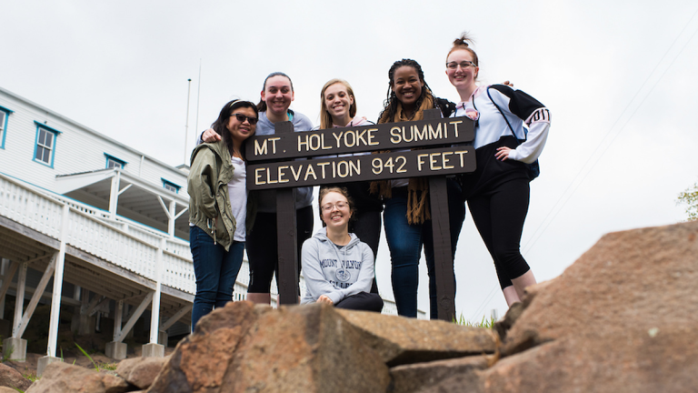 A group of students by the summit sign that says "Mt. Holyoke Summit. Elevation 942 feet"