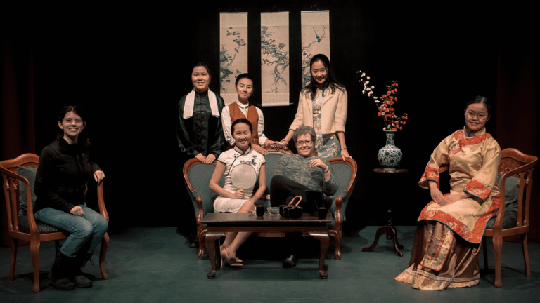 Group photo of several Asian students in traditional dress on a stage with Asian decorations and a professor.