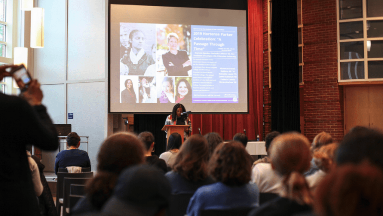 Kijua Sanders-McMurtry on stage speaking to the audience with a screen behind her and images from other Hortense Parker celebrations.