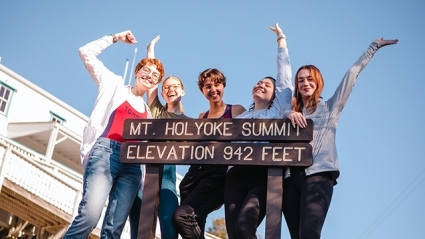 This is a photograph of students posing at the top of the mountain by a sign that reads, "Mt. Holyoke Summit, Elevation 942 Feet."