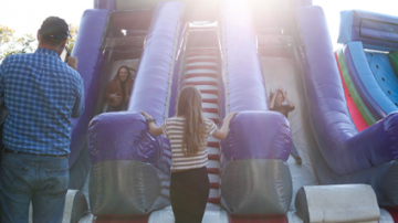 People stand at bottom of bouncy slide with the sun behind it.