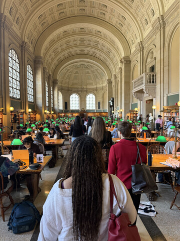 A photo of students sitting at tables, others bustling about, in a large cathedral ceiling room.