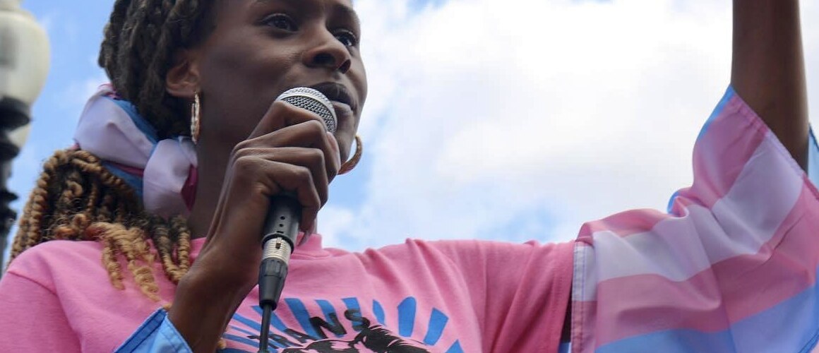 This is a photograph of Chastity Bowick at a demonstration; she is standing holding a microphone in one hand, the other arm raised. She wears a flag of pink, blue and white stripes, the colors of transgender pride. The blue sky backdrop matches the blue stripe in the flag.