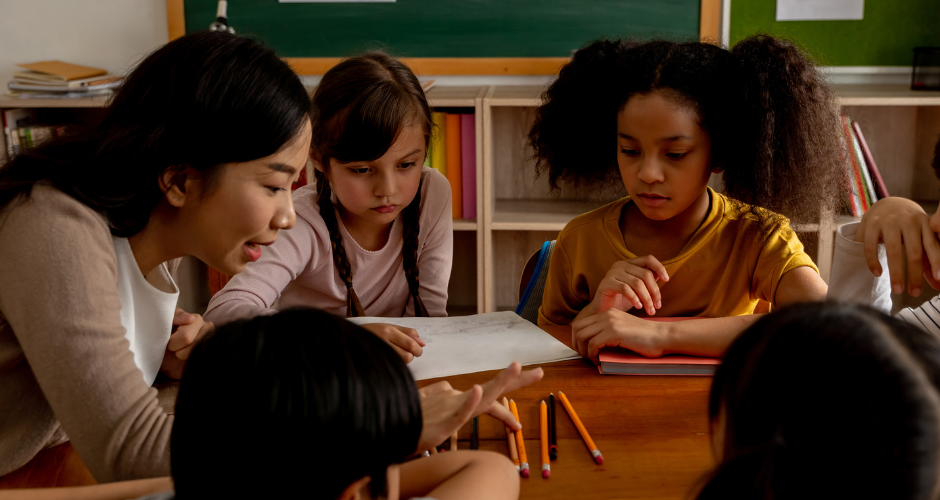 A diverse group of students are around a table, with a teacher counting pencils.