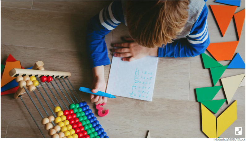 Student looking at a paper with math equations on it next to colored shapes and an abacus.